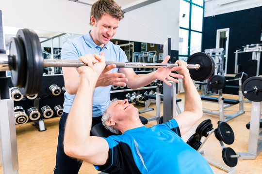 教练协助高级男子在健身房举重杠铃，以增加力量和健身