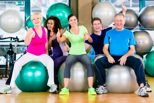 男人和女人坐在健身房的健身球上，由老人、年轻人、黑人和白人组成