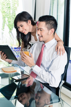 一对亚洲夫妇正在吃早餐，在这名男子上班前，他正在和妻子查看电子邮件或新闻