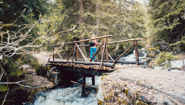 一个徒步旅行的女人站在树林里的桥上，看着山间小溪的水