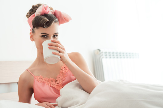 美丽的女人在床上啜饮着早晨的咖啡，开始了新的一天