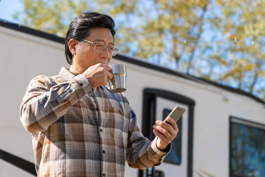 一个老年男人站在房车前看着手机视频喝咖啡