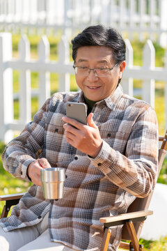 老年男人坐在露营院子里看着手机视频喝咖啡