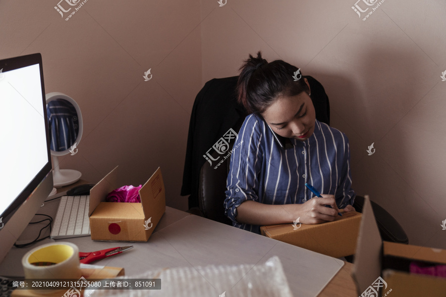 忙碌的亚泰妇女在纸板箱上写地址，和顾客通电话。复制计算机上的空间。
