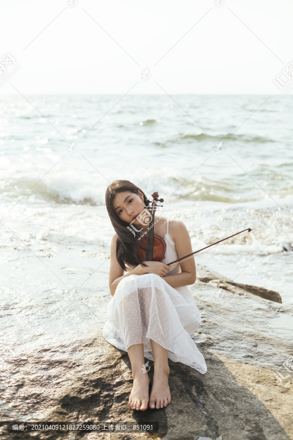 黑色长发，穿着白色连衣裙的女人，膝盖朝上，抱着小提琴坐在岩石上。