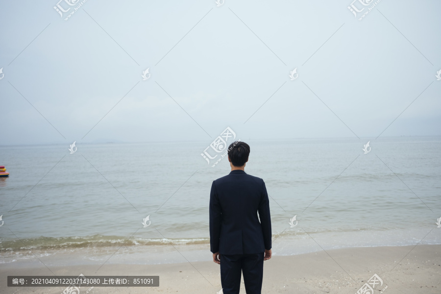 一个黑发男人，穿着深色西装，站在沙滩上，在阴暗的天空中看着大海。