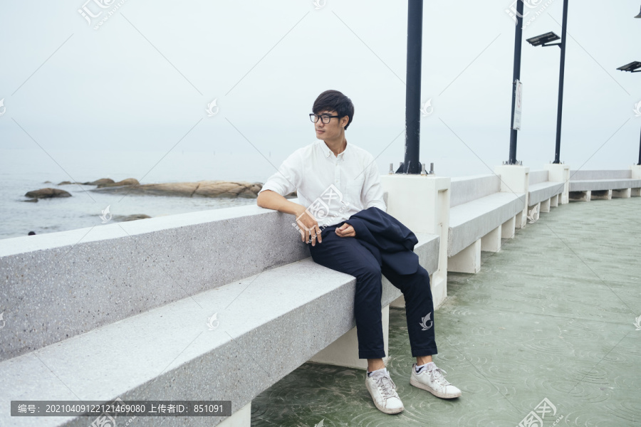 黑发汉子穿着白衬衫坐在大理石长凳上沿着码头在大海中布满了岩石。