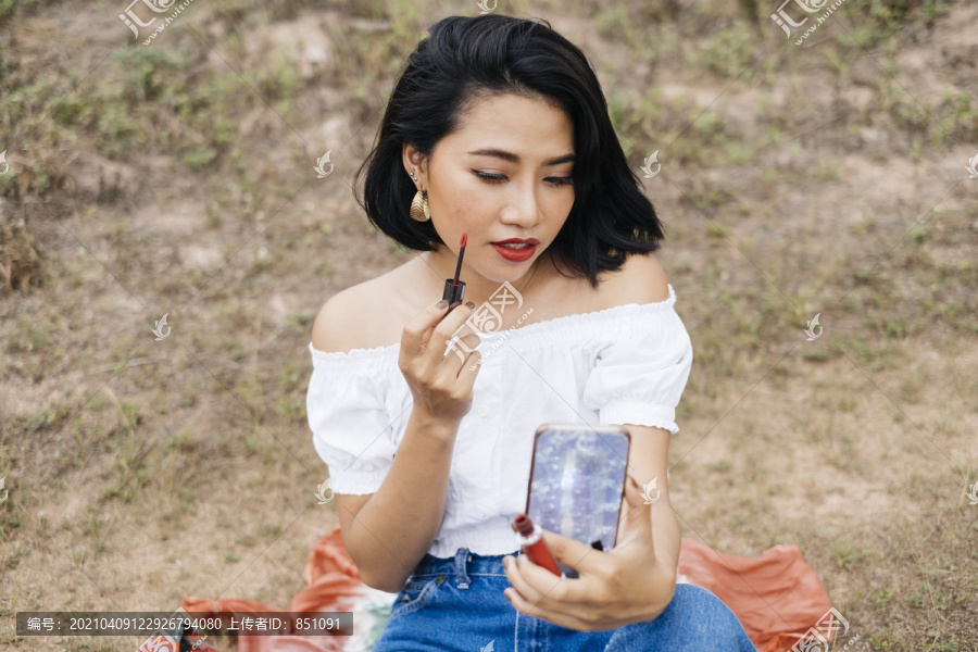 性感的亚洲年轻女人做化妆，用红色画笔涂唇膏，看着智能手机摄像头作为镜子。