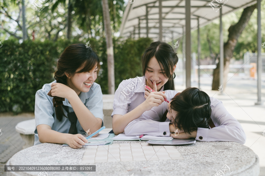 三个穿着不同制服的漂亮亚泰学生放学后一起做作业。书呆子女孩在桌子上睡着了。另外两个在恶作剧。