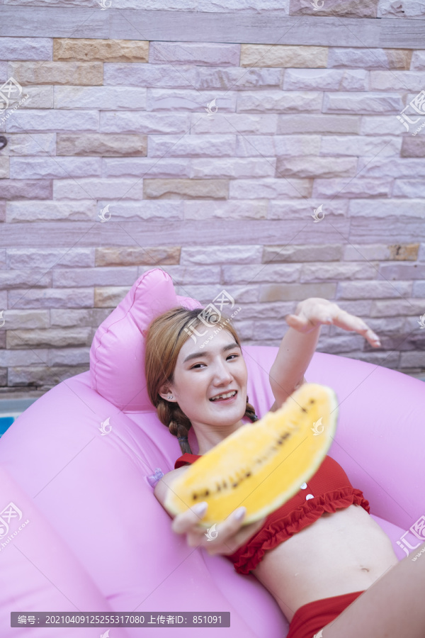 穿着红色比基尼的泰国美女躺在粉红色的泳衣上，手里拿着一块西瓜。