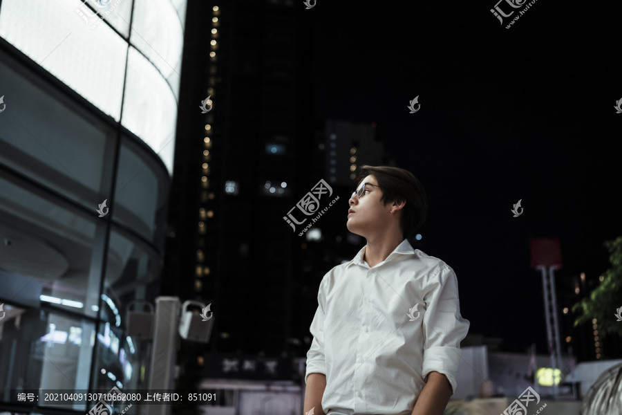 身穿白衬衫、戴着眼镜的黑发小伙儿，在夜晚从首都中心的LED广告牌旁走过购物中心。