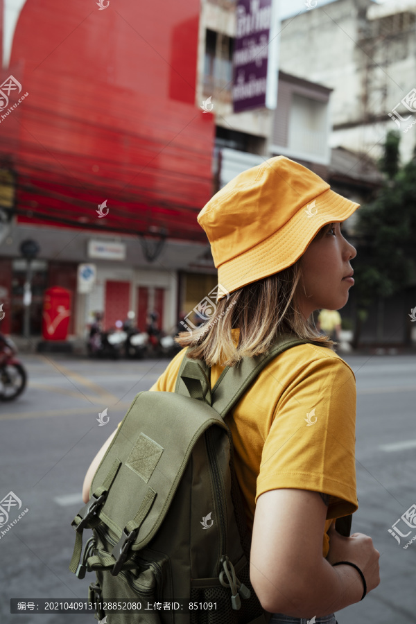 一个穿着黄色衣服，背着绿色背包的女背包客站在城市红楼对面的路边。