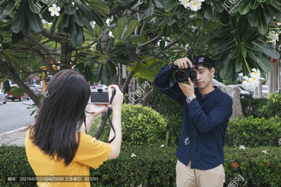 男摄影师拿着鲜花和相机，在鸡蛋花树旁为十几岁的模特女孩拍照。