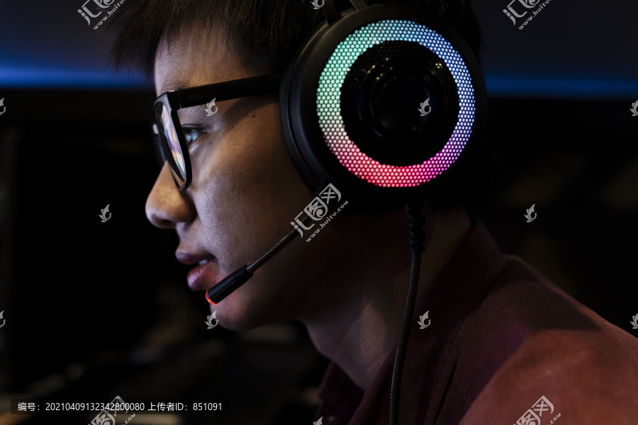特写，侧视，头像的书呆子年轻亚洲人戴着眼镜和耳机或耳机，在电脑上玩网吧，暗室。