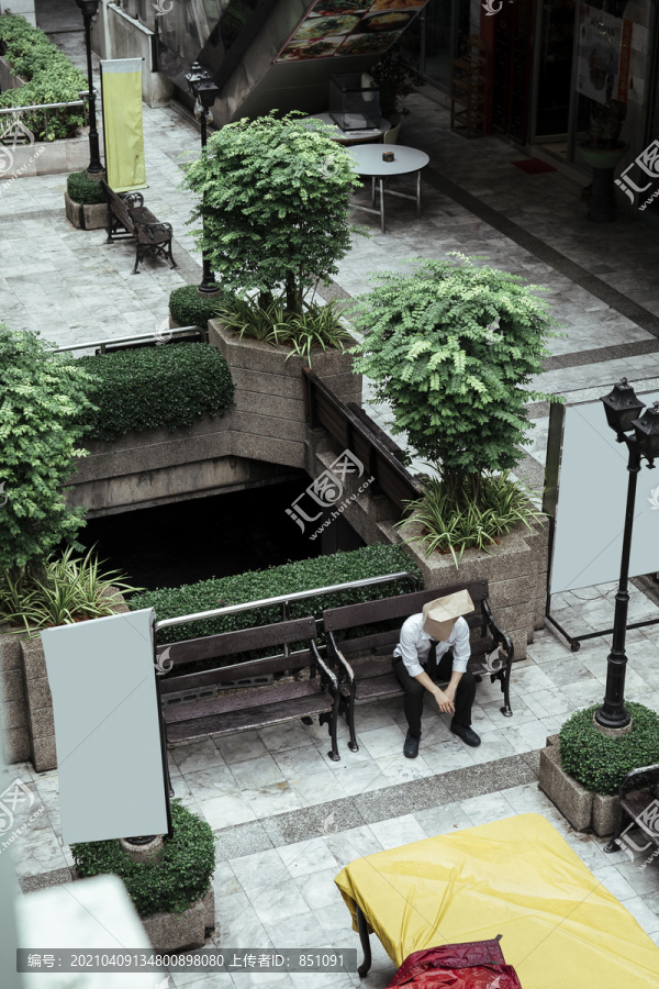 坐在城市花园广场长椅上的纸袋男。孤独的纸袋人独自坐在城市里。