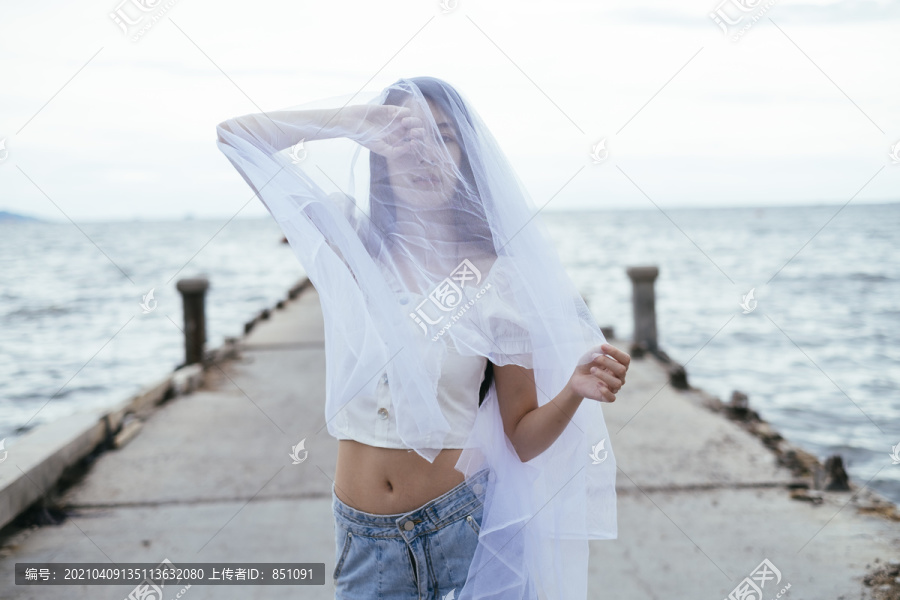 年轻的亚洲长发黑发女子戴着白纱在海桥上的肖像。衬衫和蓝色牛仔裤长裤。