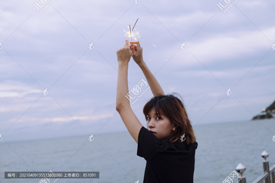 背景图：年轻的泰国亚裔女子短发举手举着两个火花头在海滩上在杜什时间。回头看摄像机。