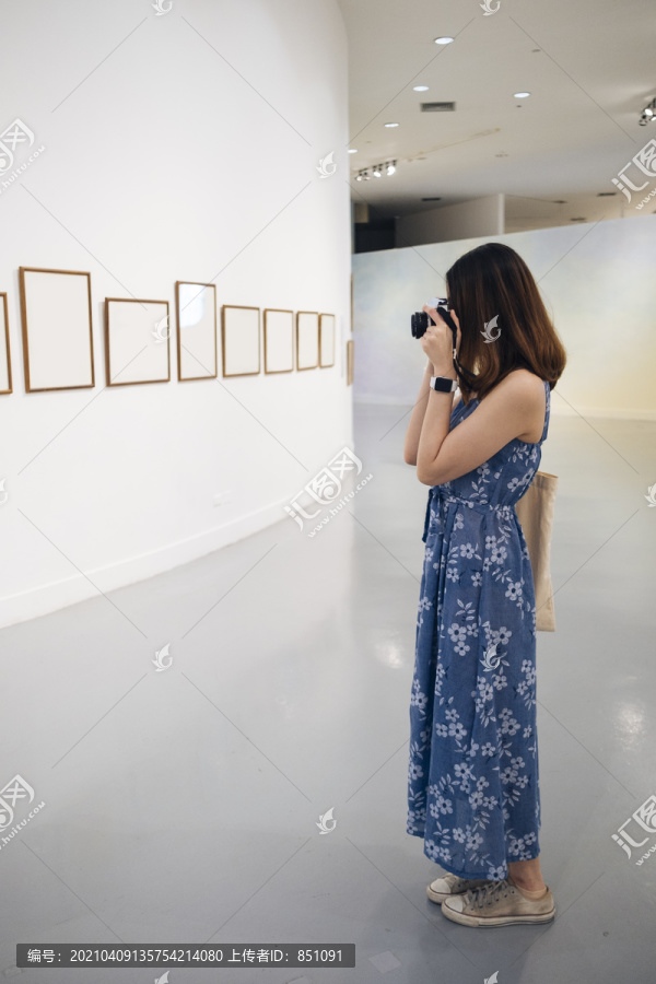 在美术馆里，一位身着蓝色连衣裙的泰国亚裔年轻女子正在用相机拍照。