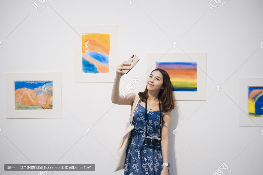 在美术馆里，一位穿着蓝色裙子的亚洲时髦美女正在用智能手机自拍。