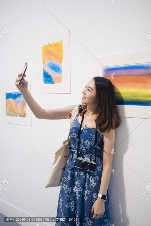 在美术馆里，一位穿着蓝色裙子的亚洲时髦美女正在用智能手机自拍。