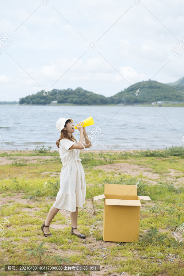 在湖边的纸板箱旁，一位年轻的泰国亚裔女子卷发，身着白色连衣裙，玩着黄色玩具扬声器扩音器。