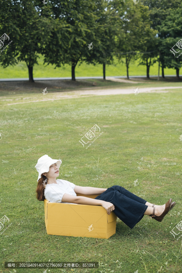 年轻的泰国亚裔女子嬉皮卷发，穿着白衬衫，躺在绿色公园草地上的纸板箱上。