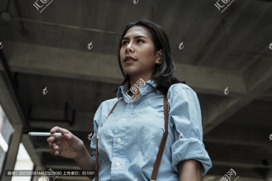 身着蓝色衬衫的亚泰妇女手持香烟在地下室散步的肖像。