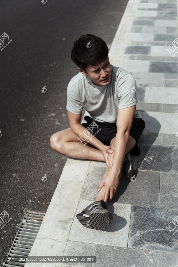 一个亚裔运动员坐在路边腿抽筋，表情很难看。健康问题概念。腿抽筋引起的疼痛。