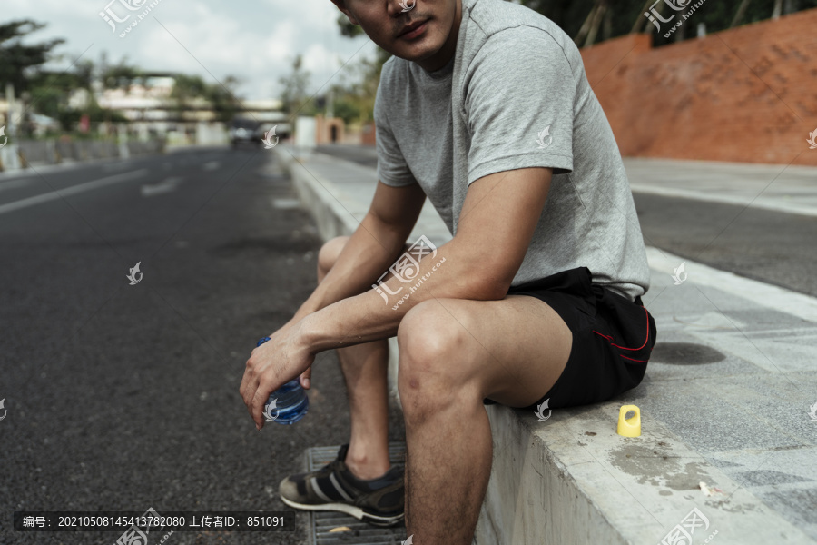 坐在人行道上拿着一瓶水的亚洲人。一个男人拿着一瓶水，坐在路边。
