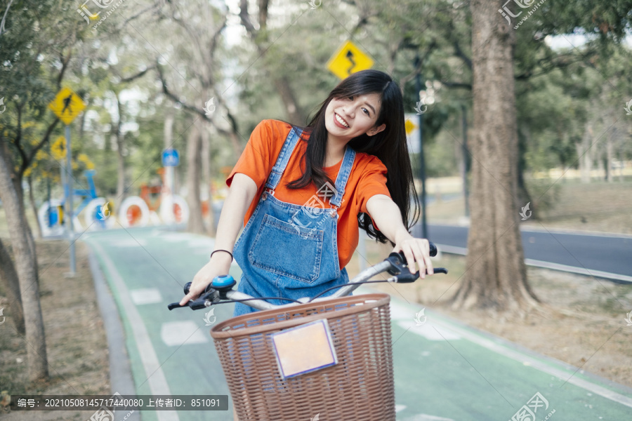 长发女孩穿着橙色t恤和牛仔裤在公园的自行车道上骑自行车，靠近镜头。