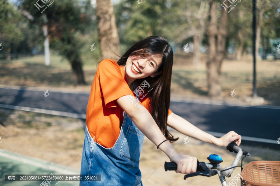 长发女孩穿着橙色t恤和牛仔裤，在公园的自行车道上停下自行车，然后歪着头看着镜头，微笑着。