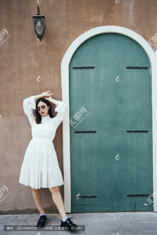 美丽的白衣长发女孩站在绿色门和灯的棕色墙前，姿态别致。