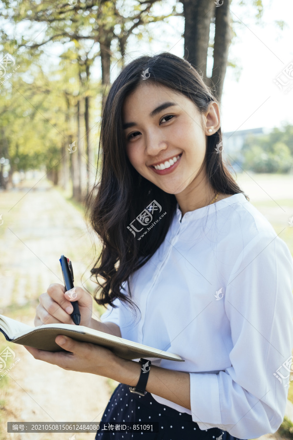 快乐的亚洲女人穿着白衬衫写着一本书，带着快乐的感觉，笑着笑着。小说作家微笑着写作的女孩。