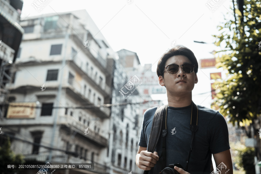 年轻英俊的亚洲摄影师戴着太阳镜拿着相机走在曼谷街头。
