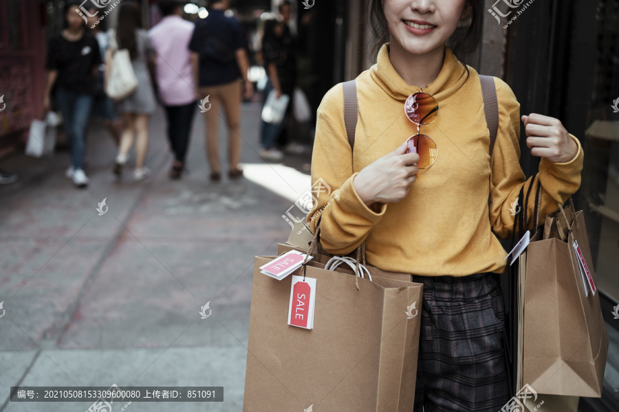 美丽的亚洲女人穿着黄色毛衣，背着许多贴着价签的购物袋，走在街上。