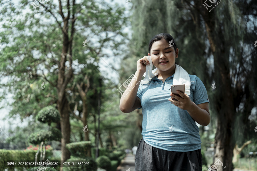 疲惫胖胖的运动型亚洲女性，穿着蓝色运动服，满头大汗，白毛巾，看着智能手机，查看网站，和朋友聊天。