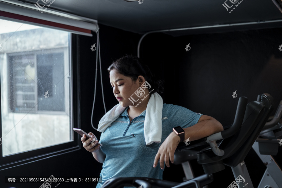 胖乎乎的运动型亚洲女性，穿着蓝色运动服，厌倦了在健身房里用智能手机锻炼。