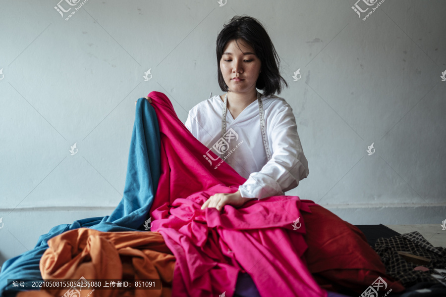 一位年轻的亚洲设计师，一头短发，穿着白衬衫，从一堆布料中挑选布料来做衣服。