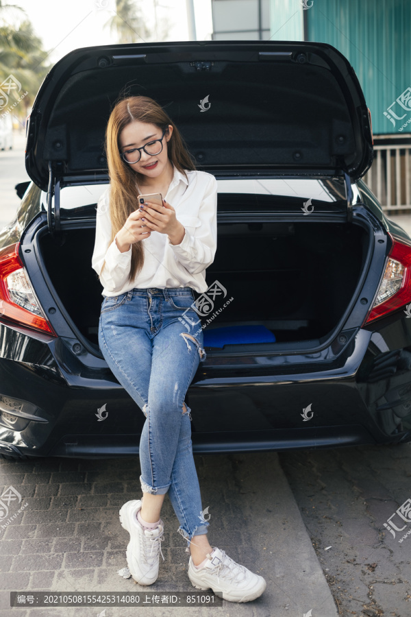 交通和车辆概念——戴眼镜的泰国妇女坐在汽车后备箱旁，通过智能手机与人聊天发短信。