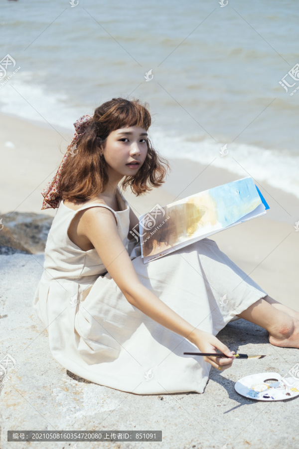 一幅美丽的亚泰艺术家的肖像画，身着白色连衣裙，坐在沙滩上，用小毛笔画上带有液体色彩的洞穴。