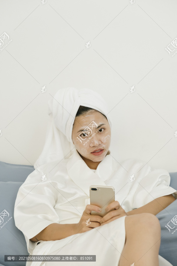 穿着白色浴袍的年轻女子用智能手机敷化妆面膜，躺在卧室的床上和人们在社交网络上聊天。