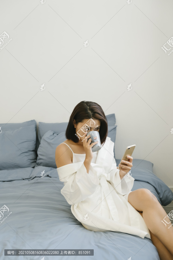 穿着白衬衫的性感亚泰女人坐在床上，拿着智能手机，从马克杯里喝着热茶和咖啡。