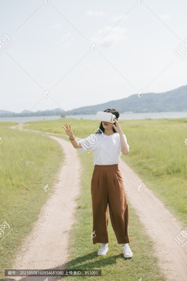 黑色长发女子穿着休闲服，戴着VR护目镜，在泥泞的草原上行走，享受虚拟现实技术。