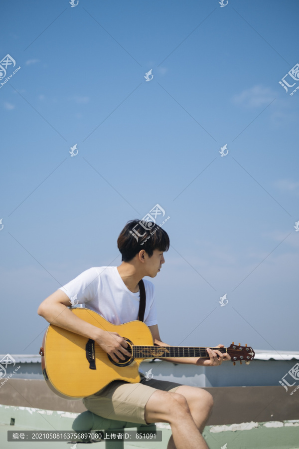 在晴朗的天空下，一个年轻的泰国吉他手坐在一个管子上，在屋顶上弹原声吉他。