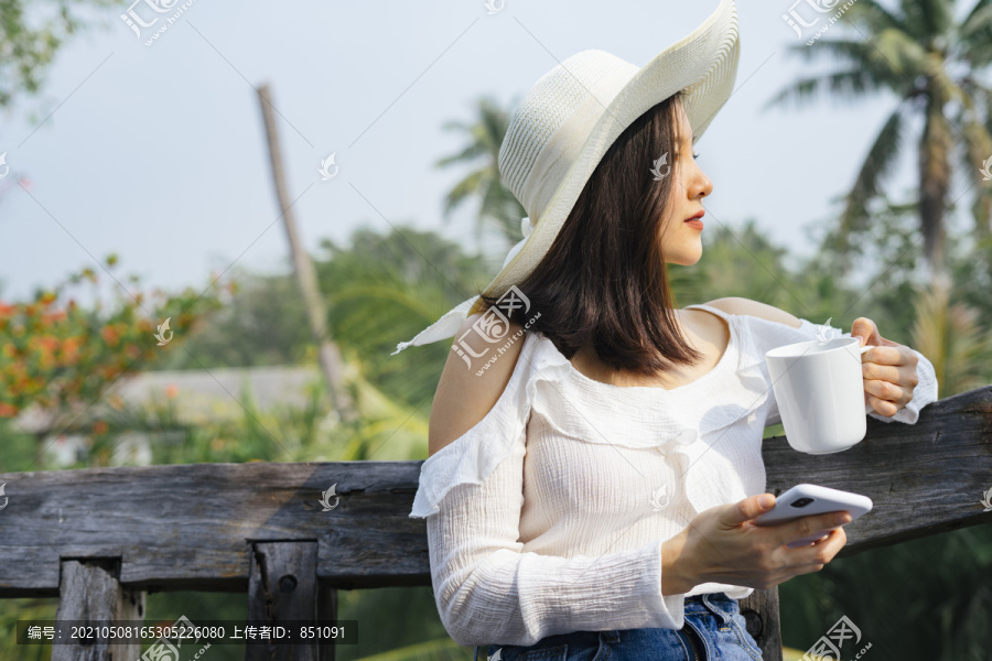 一位身穿白衬衫的亚泰妇女端着一杯咖啡，坐在木凳上，用智能手机与朋友联系。
