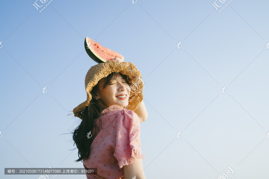 戴着棕色帽子的可爱女孩抬起头上的西瓜，在蓝天下微笑。