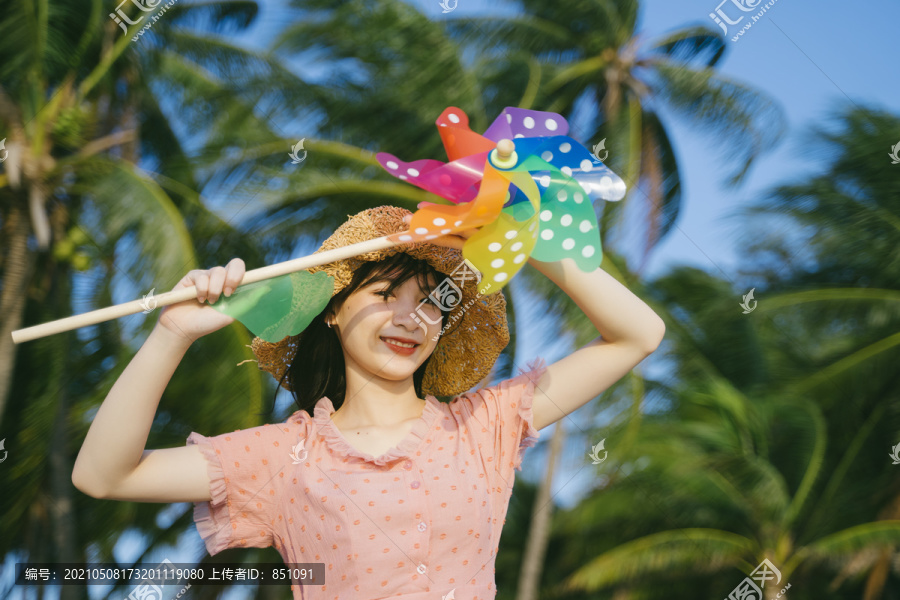 风和日丽的沙滩上，戴着棕色帽子的可爱女孩手持五颜六色的风车在椰子树前。