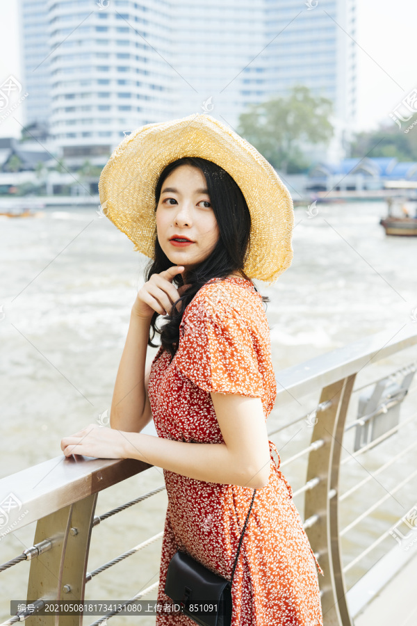 年轻漂亮的泰国亚裔旅行家，穿着红色连衣裙，戴着帽子，站在曼谷朝普拉亚河的码头上。