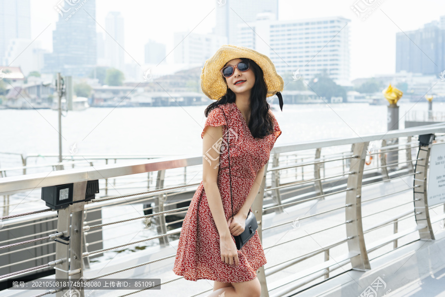 一位年轻漂亮的泰国亚裔旅行家，穿着红色连衣裙，戴着帽子，戴着太阳镜，在曼谷市的一个码头上旅行。