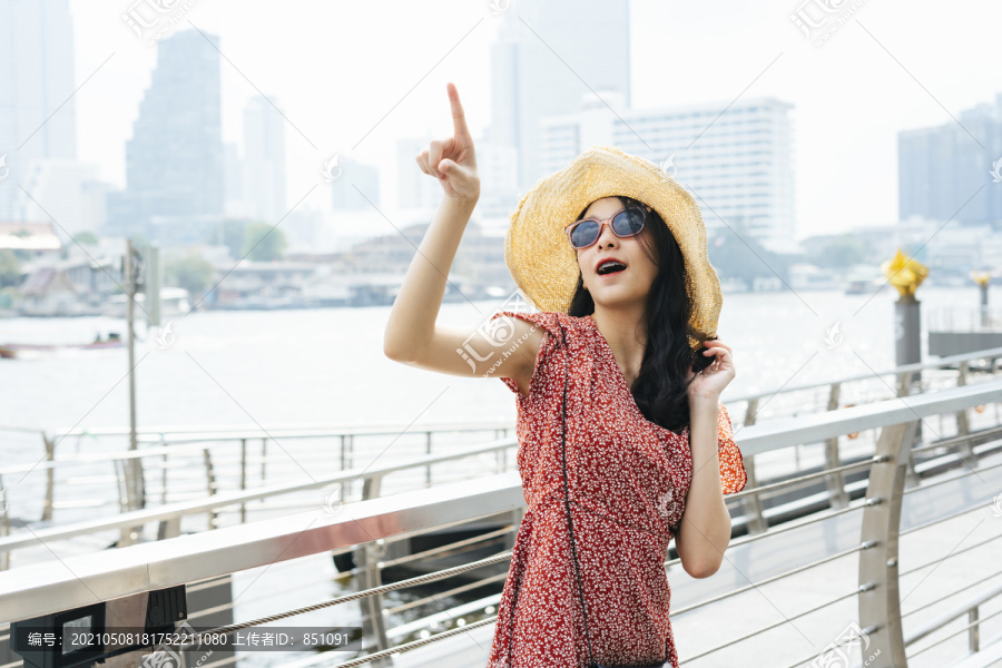 年轻漂亮的泰国亚裔旅行家，穿着红色连衣裙，戴着帽子，戴着太阳镜，指着天空。曼谷市的一位女旅行者。